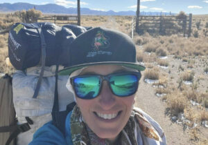 a woman hiker on a dirt road in front of an old western corral with mountains in the background